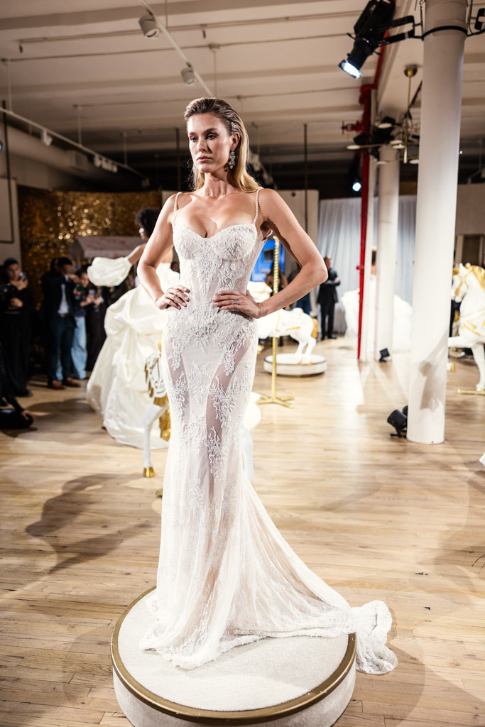 Woman in a fitted, lace white gown poses confidently on a round platform at an indoor New York Bridal Fashion Week event.