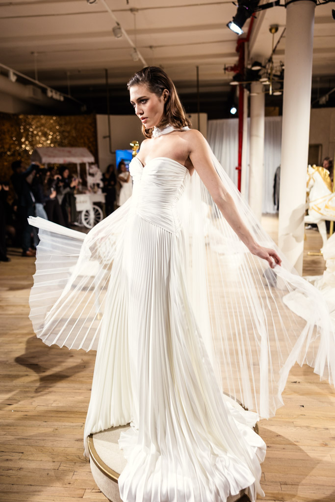 A woman in a strapless, pleated white gown poses indoors on a small platform, holding out the fabric—capturing the elegance of New York Bridal Fashion Week.