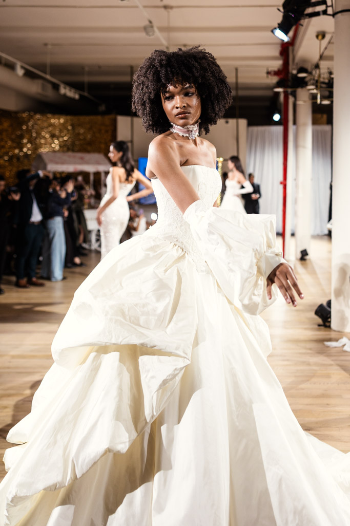 A model in a dramatic white gown walks the runway at New York Bridal Fashion Week, with onlookers in the background.