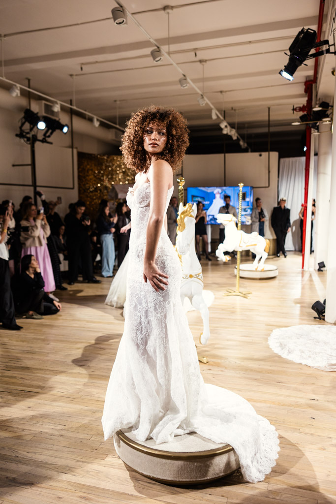 A model in a white lace wedding dress poses on a platform at an indoor fashion event during New York Bridal Fashion Week, captivating the onlookers.