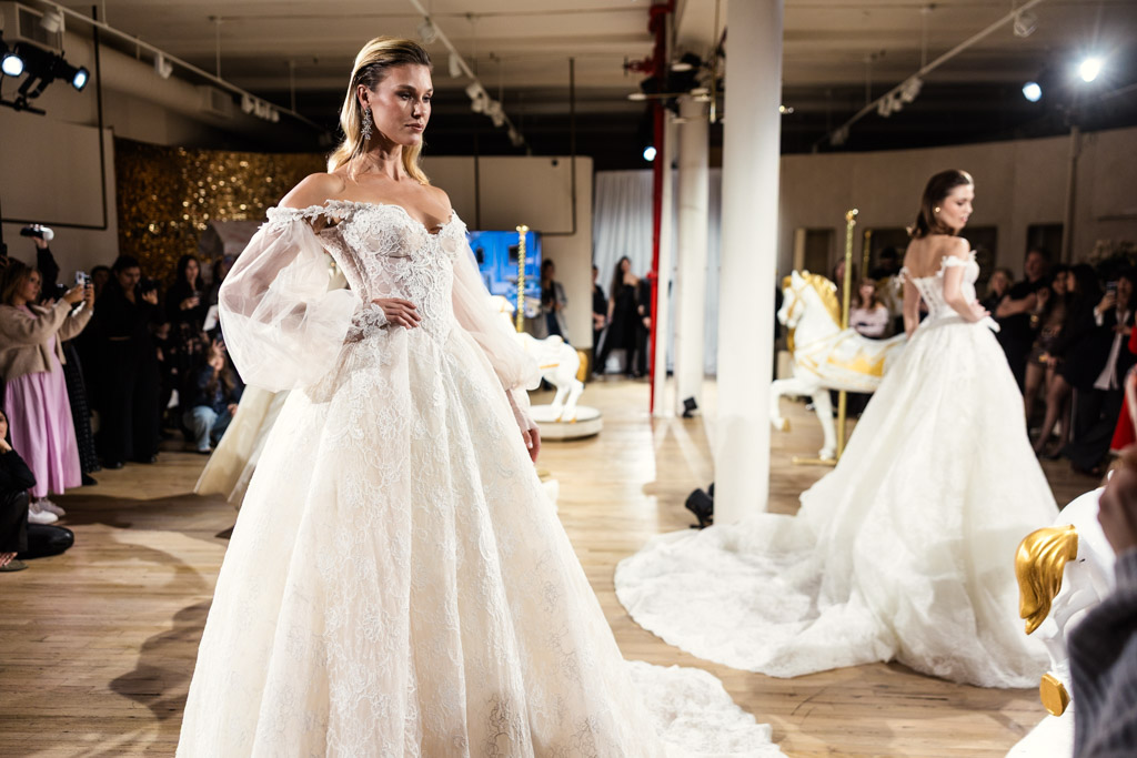 Model in an elegant off-shoulder wedding gown walking a runway at New York Bridal Fashion Week, with another model and carousel horses behind.