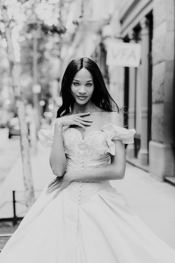 A woman in an elegant gown poses confidently on a city sidewalk, capturing the chic atmosphere of New York Bridal Fashion Week with stunning buildings in the background.