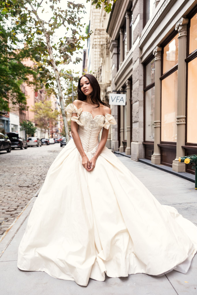 Woman in an off-the-shoulder, cream ball gown standing on a city sidewalk, reminiscent of New York Bridal Fashion Week elegance, with trees and buildings in the background.