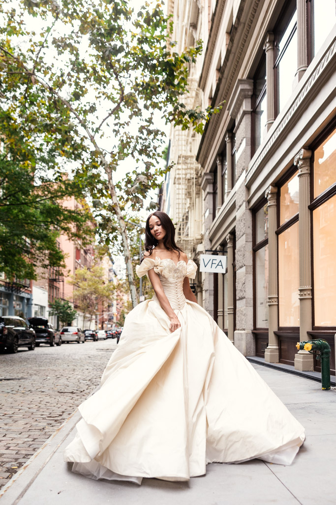 Woman in an elegant off-shoulder ball gown posing on a city sidewalk near buildings and trees, capturing the glamour of New York Bridal Fashion Week.