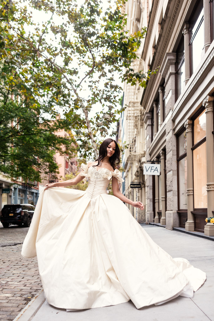 A woman in an elegant white gown poses on a city sidewalk near historic buildings and trees, capturing the timeless glamour of New York Bridal Fashion Week.
