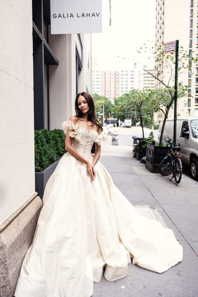 A woman in an elegant, off-shoulder ivory gown stands outside a Galia Lahav boutique on a city sidewalk during New York Bridal Fashion Week.