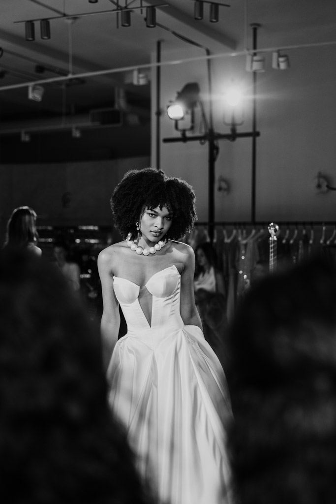 A model in a strapless gown walks the runway at New York Bridal Fashion Week, with lights and racks in the background.