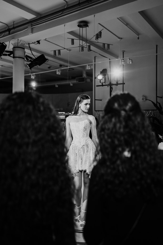 A model in a lacy dress walks the runway at New York Bridal Fashion Week, viewed from behind curly-haired audience members.