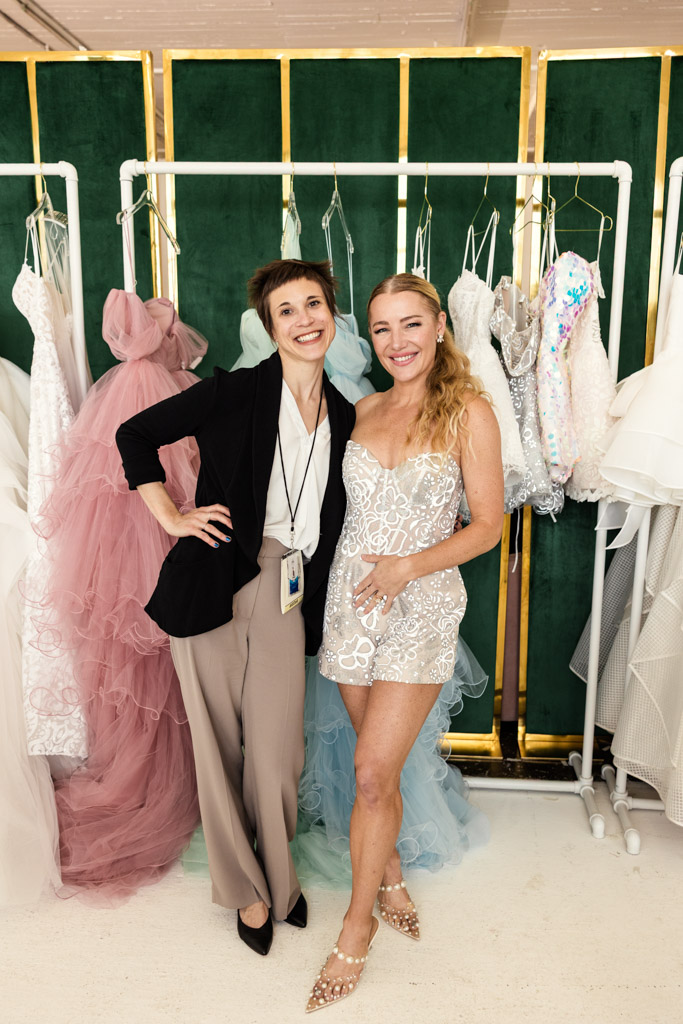 Two women posing and smiling in front of racks of elegant, pastel-colored dresses at New York Bridal Fashion Week.