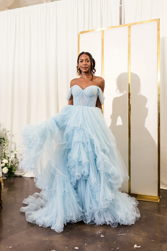 A woman in a flowing light blue off-the-shoulder gown stands indoors before cream curtains and a gold screen, embodying the elegance seen at New York Bridal Fashion Week.