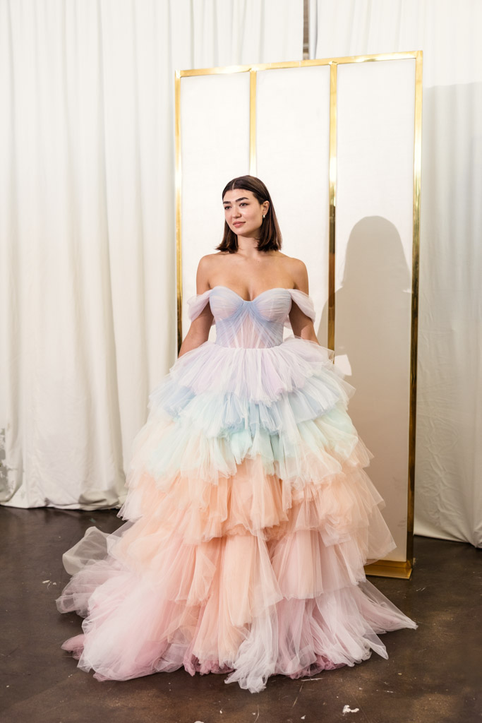A woman in a pastel, layered tulle gown stands indoors before white curtains and a gold-framed screen, embodying elegance inspired by New York Bridal Fashion Week.