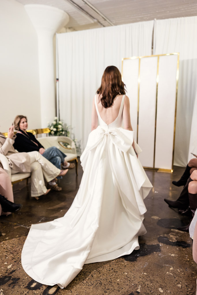 A bride in a white gown with a large bow on the back walks down an aisle, capturing the elegance of New York Bridal Fashion Week as seated guests watch.