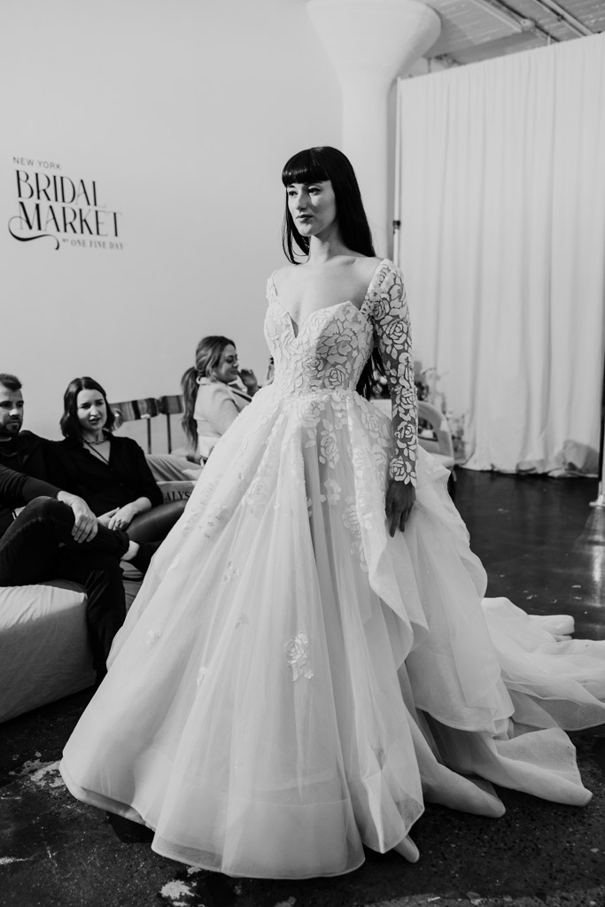 A model in a long-sleeve lace wedding dress walks the runway at New York Bridal Fashion Week, with seated guests watching.