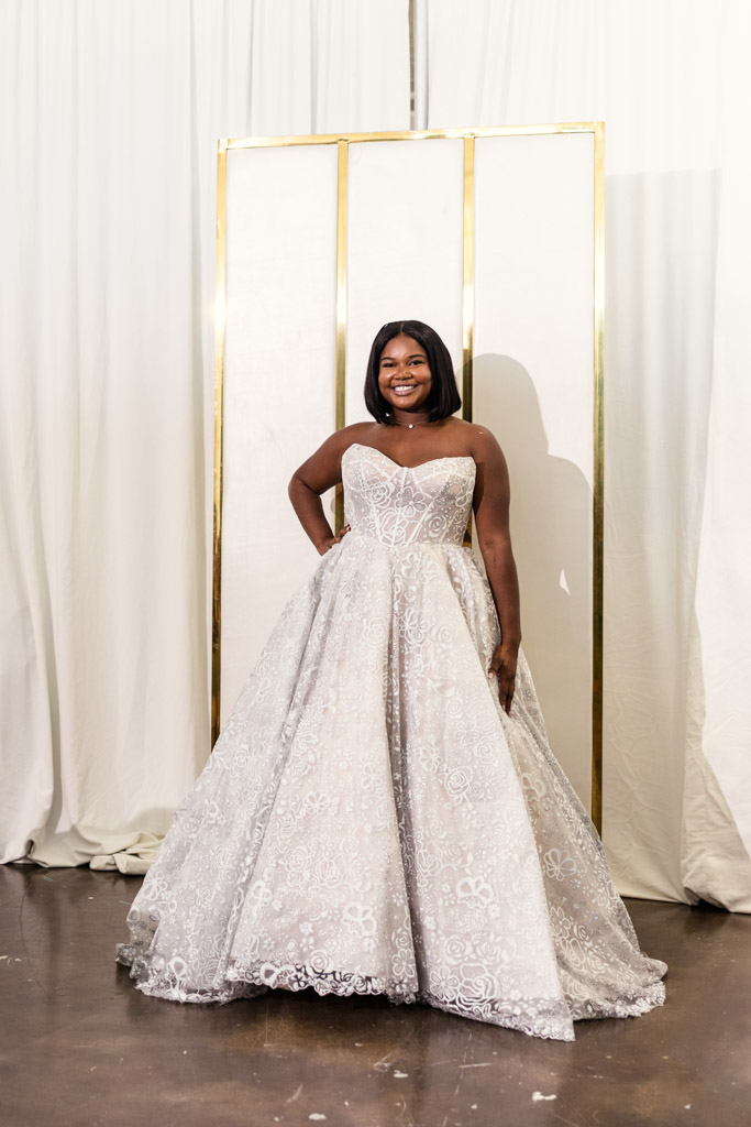 Smiling woman in a strapless, lacy ballgown stands in front of a white and gold backdrop at New York Bridal Fashion Week.