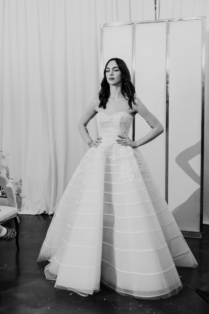 A woman stands with hands on hips, wearing a strapless, full-skirted wedding gown in a bright room, reminiscent of styles seen at New York Bridal Fashion Week.