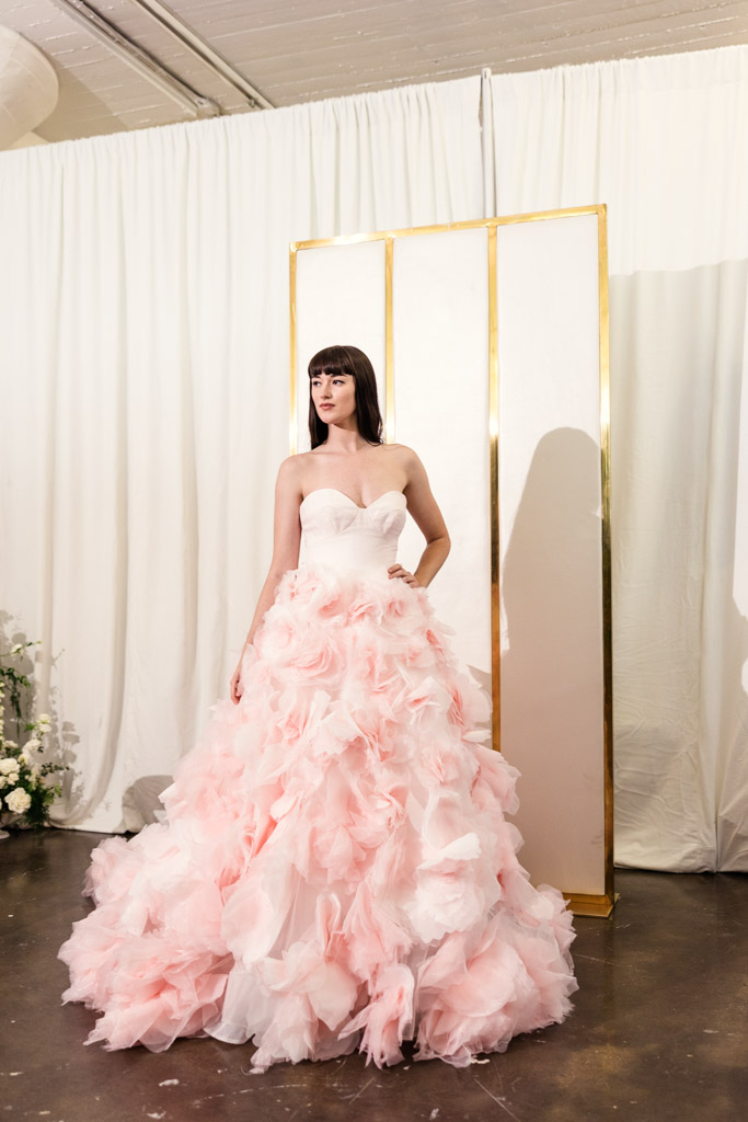 A woman in a strapless white dress with a voluminous, fluffy pink skirt stands in front of white curtains, evoking the glamorous style seen at New York Bridal Fashion Week.