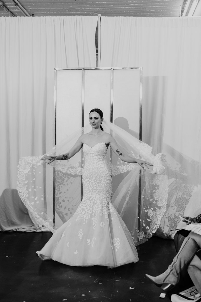 A bride in a strapless, lace mermaid gown poses, holding out a long, floral lace veil in front of white curtains—a timeless look inspired by New York Bridal Fashion Week.