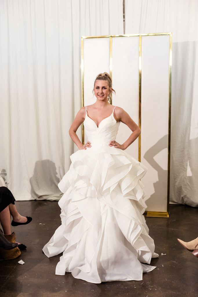 Woman in a ruffled white wedding gown stands smiling with hands on hips, in front of white curtains, capturing the elegance seen at New York Bridal Fashion Week.