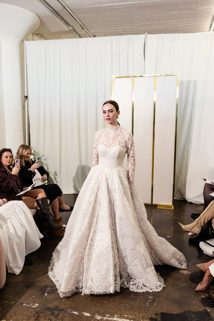 A model in a lace wedding gown walks the runway at New York Bridal Fashion Week, surrounded by seated spectators in a white room.