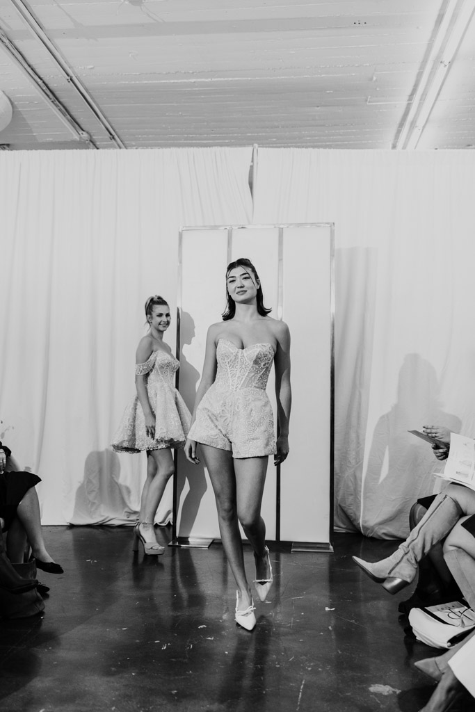 Two models walk the runway in short, elegant dresses at a New York Bridal Fashion Week show, with audience members seated nearby.