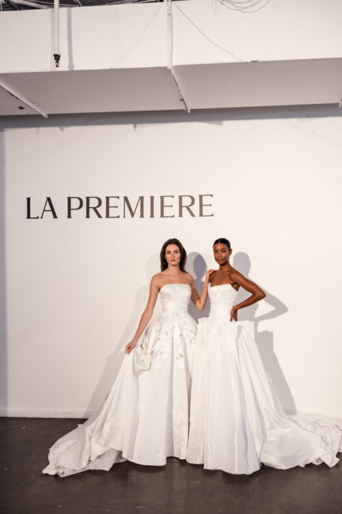 Two women wearing elegant white wedding gowns stand in front of a wall that reads "LA PREMIERE," capturing the chic atmosphere of New York Bridal Fashion Week.