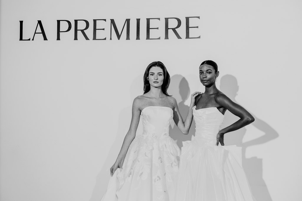 Two women in elegant white gowns pose under a sign that reads