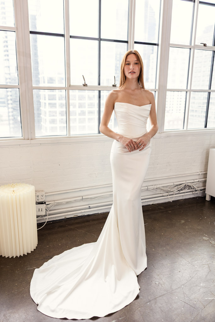 A woman in a strapless white wedding gown stands by large windows in a bright, modern room, capturing the elegance showcased at New York Bridal Fashion Week.
