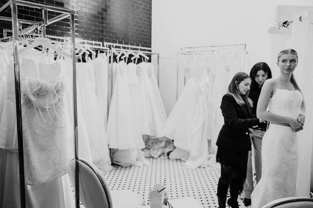A woman tries on a wedding dress with the help of two others in a boutique filled with gowns, inspired by styles from New York Bridal Fashion Week.