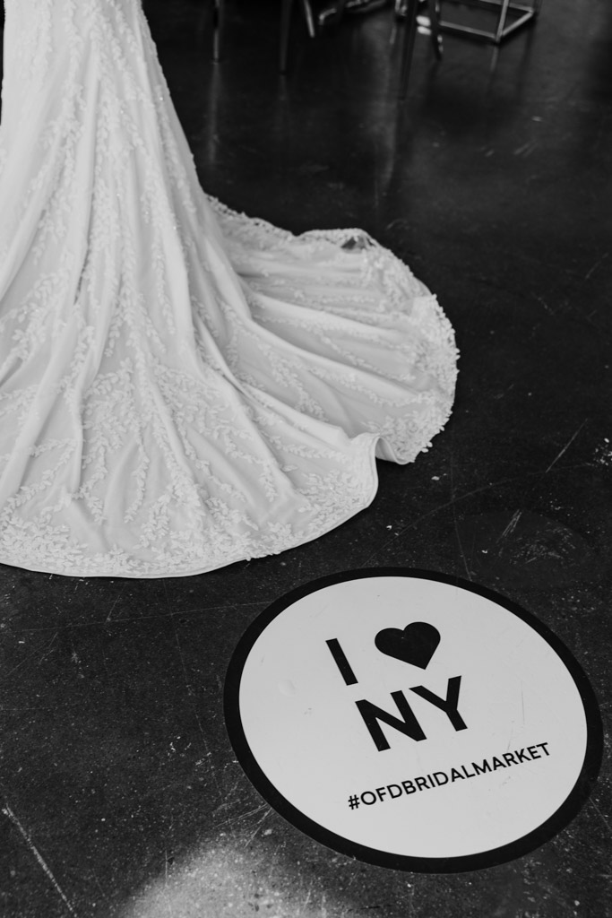Black-and-white photo of a bridal gown train near a round “I ♥ NY” #OFDBRIDALMARKET floor sign, capturing the elegance and excitement of New York Bridal Fashion Week.