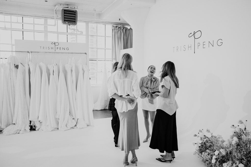 Four women stand talking and laughing in a bridal boutique adorned with wedding dresses and floral decor, inspired by the latest styles from New York Bridal Fashion Week.