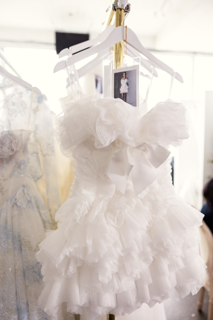 Short white ruffled dress with a large bow on a hanger, displayed next to another dress in a bright room—capturing the elegance of New York Bridal Fashion Week.