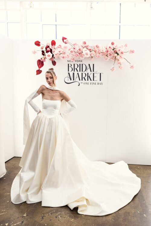 Bride in elegant white gown poses under "New York Bridal Market" sign with pink flower decor at New York Bridal Fashion Week.