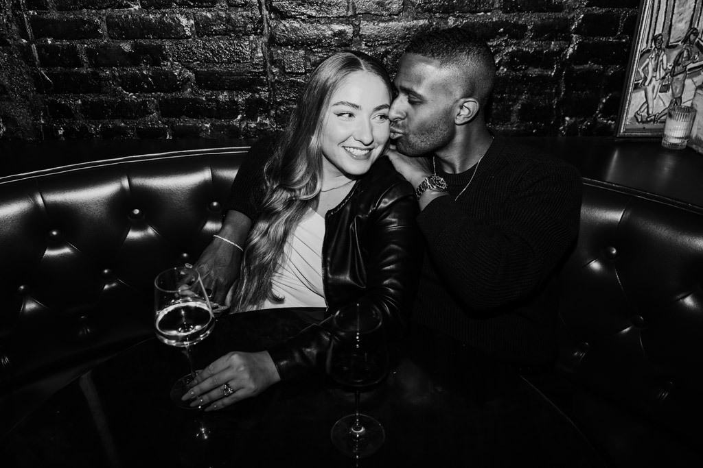 Black and white photo of man kissing his partner's cheek as they sit in a booth at Emilio's Ballato during New York City engagement session
