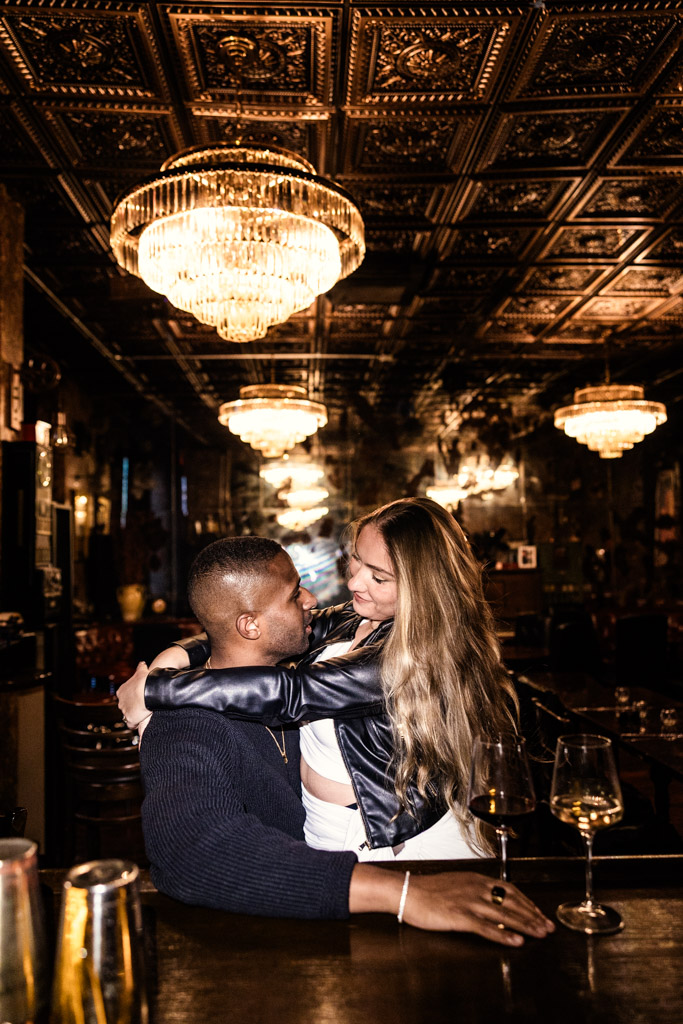 A couple embraces and smiles at each other during their NYC engagement session at the bar inside Emilio's Ballato with chandeliers and ornate ceiling decor