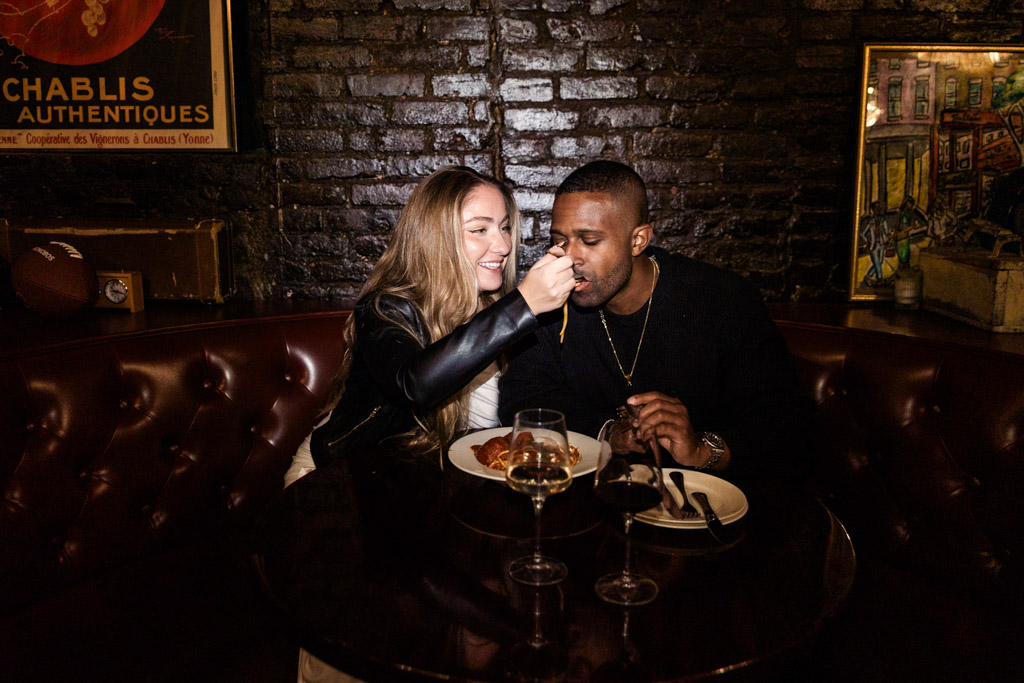 Woman feeds a man pasta at a cozy restaurant table inside Emilio's Ballato during their New York City engagement session