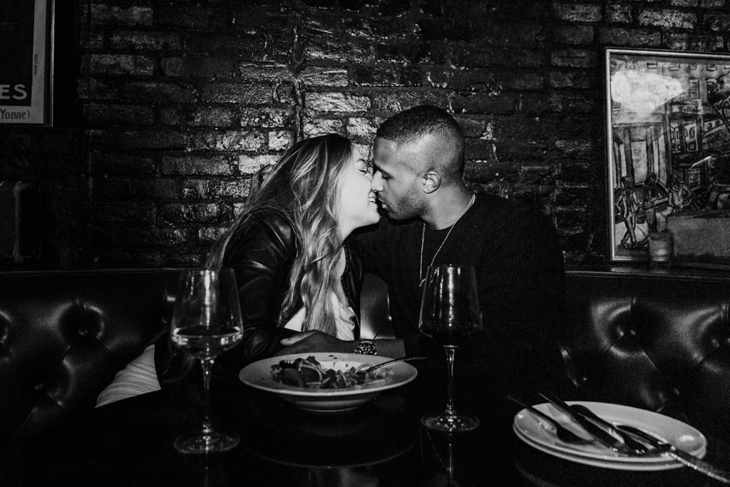 Black and white photo of engaged couple leaning in for a kiss as they sit in a booth at Emilio's Ballato during their NYC engagement session