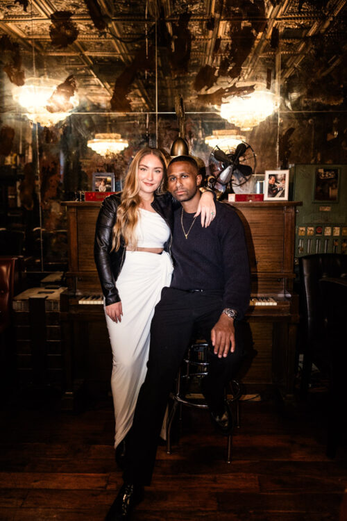 Portrait of woman in a white dress standing beside a man in black, seated by a piano inside Emilio's Ballato in New York City during their fall engagement session