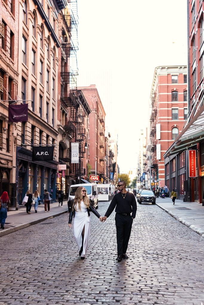 A couple holding hands walks down a cobblestone street lined with buildings in New York City during their fall engagement session
