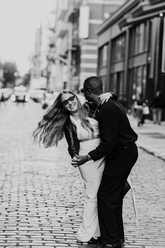 Black and white photo of engaged couple laughing and dancing together on a cobblestone New York City street