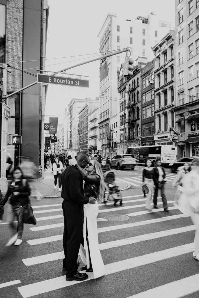 Creative black and white motion-blur photo of couple holding each other in the middle of a busy E Houston St crosswalk as people walk around them during their New York City engagement session
