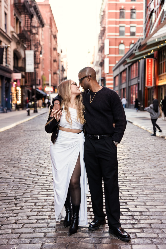 Couple stands close together, holding hands and smiling on a cobblestone street during their New York City engagement session