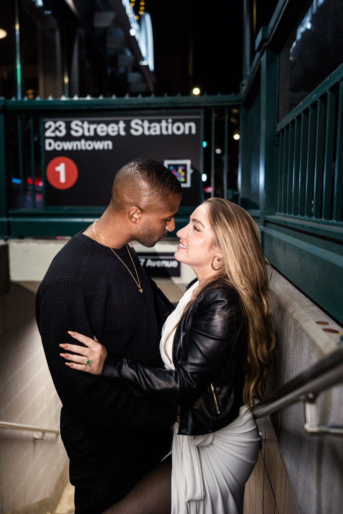 Engaged couple smiles standing close together by the 23 Street Station subway entrance at night