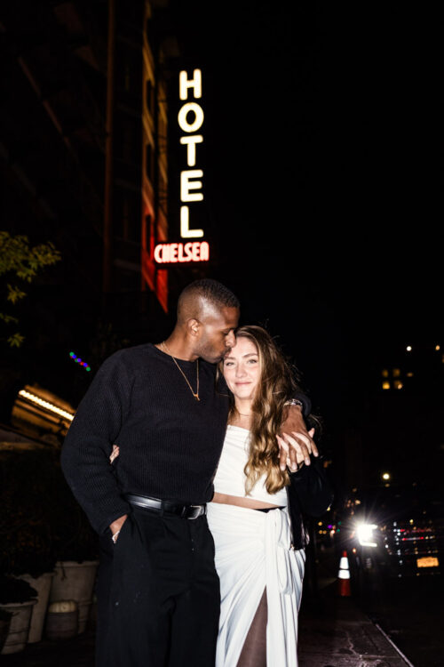 A couple stands close at night under the neon Hotel Chelsea sign, city lights glowing in the background during their New York City engagement session