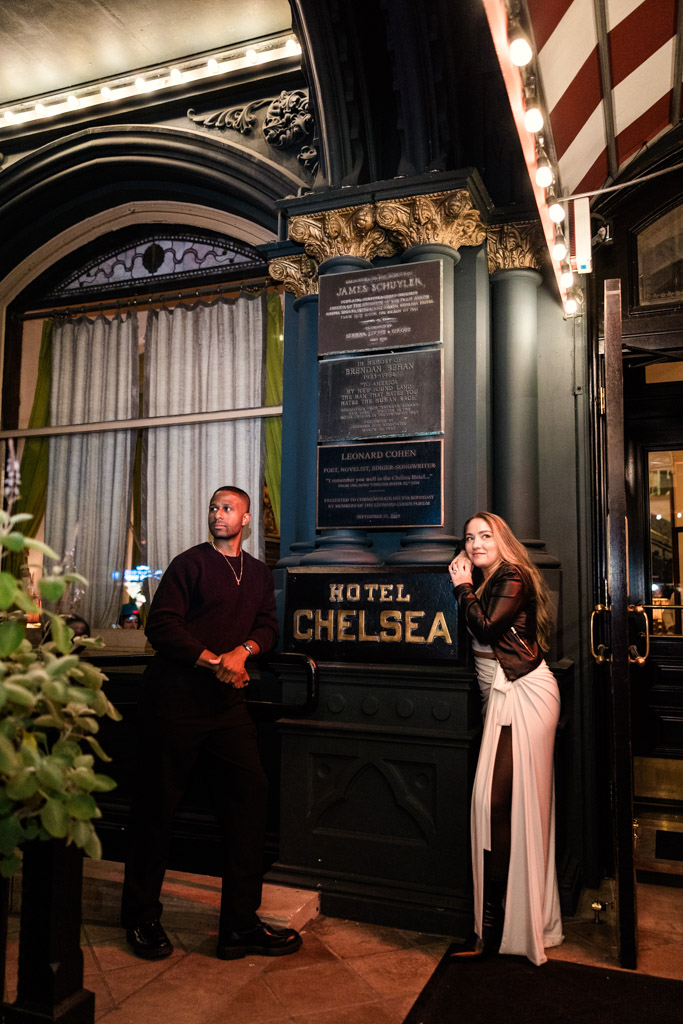 Man and woman pose outside the entrance of Hotel Chelsea at night during their NYC engagement session