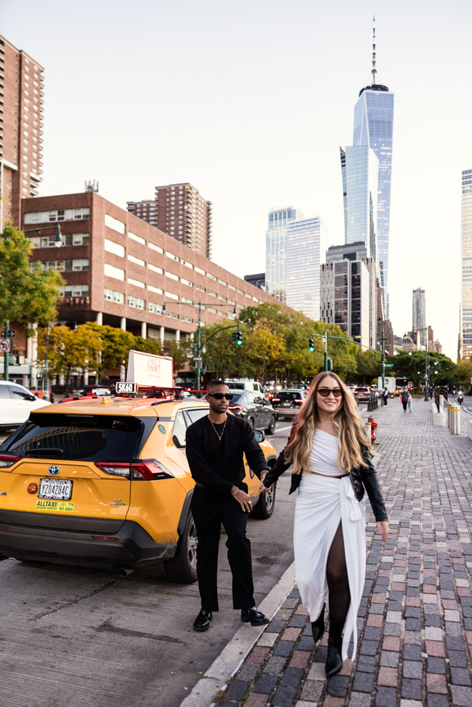 A stylish couple walks on a cobblestone sidewalk near a yellow taxi, with skyscrapers in the background during their New York City engagement session
