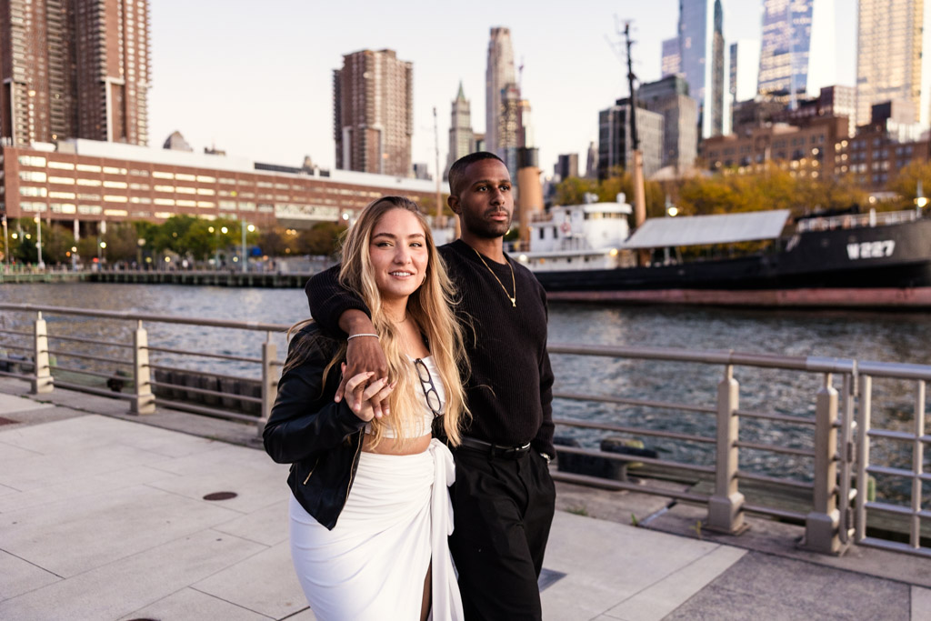 Couple stands by a riverside railing in Hudson River Park with city buildings and a docked ship in the background