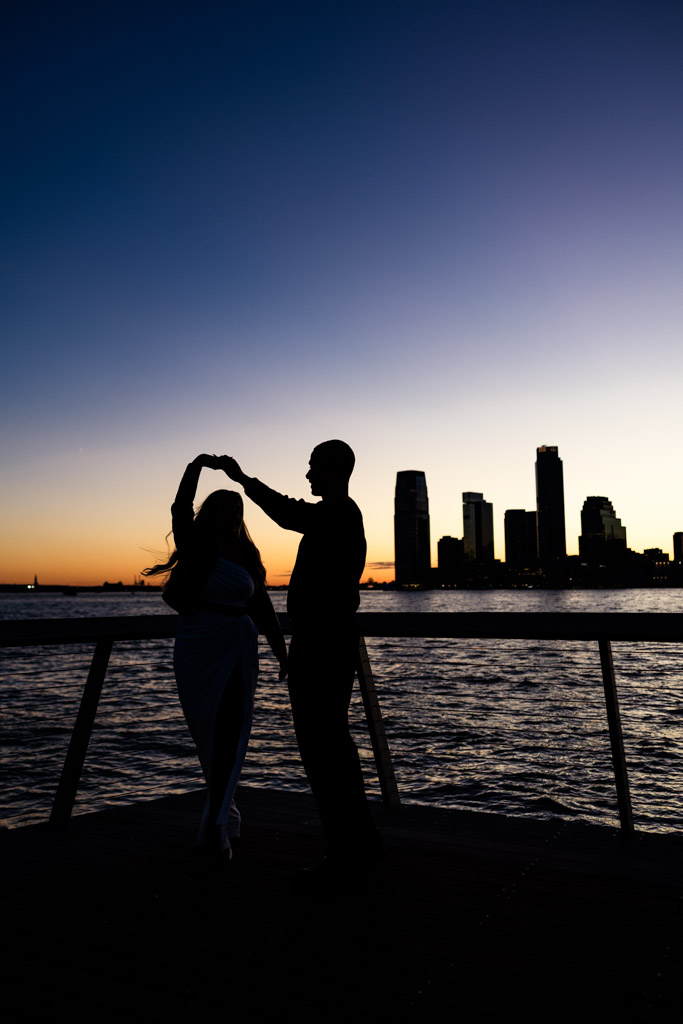 Silhouette of engaged couple dancing by the water at sunset, with the NYC skyline in the background