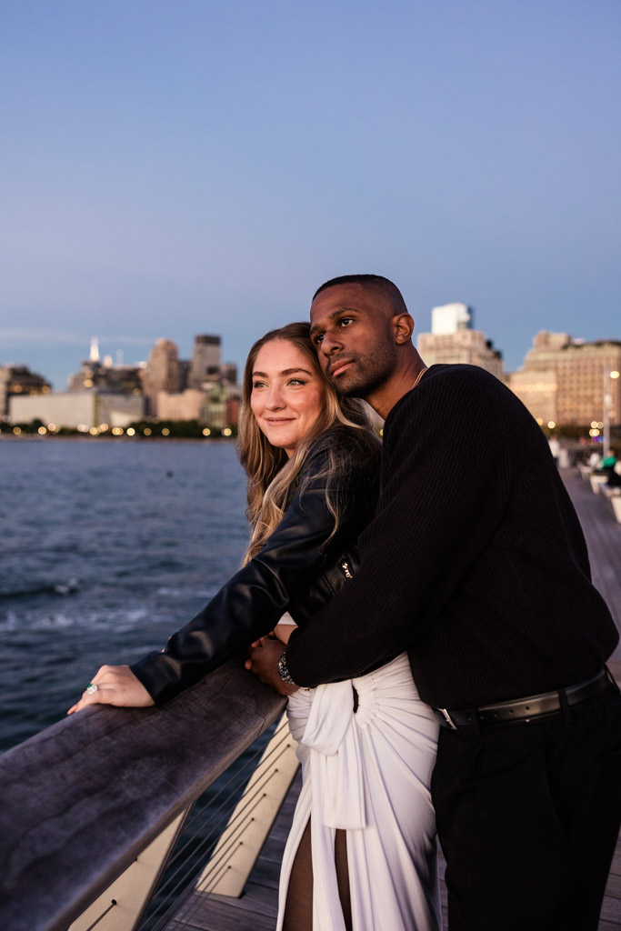 Engaged couple stands by a Hudson River Park waterfront railing at sunset, the city skyline glowing behind them