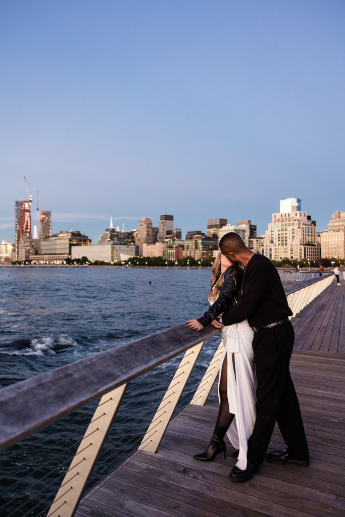 Engaged couple kisses on a Hudson River City boardwalk by the water during sunset, with a city skyline in the background