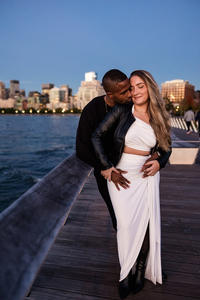 Romantic photo of engaged couple embracing on a Hudson River Park boardwalk by the water at sunset with the New York City skyline in the background